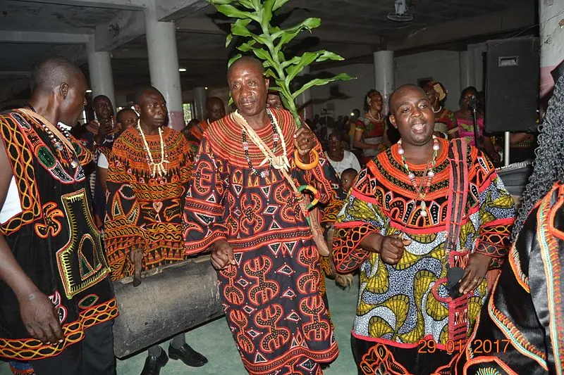 Dancers in embroidered toghu robes performing the Njang, one carrying a leafy branch, a wooden drum behind them