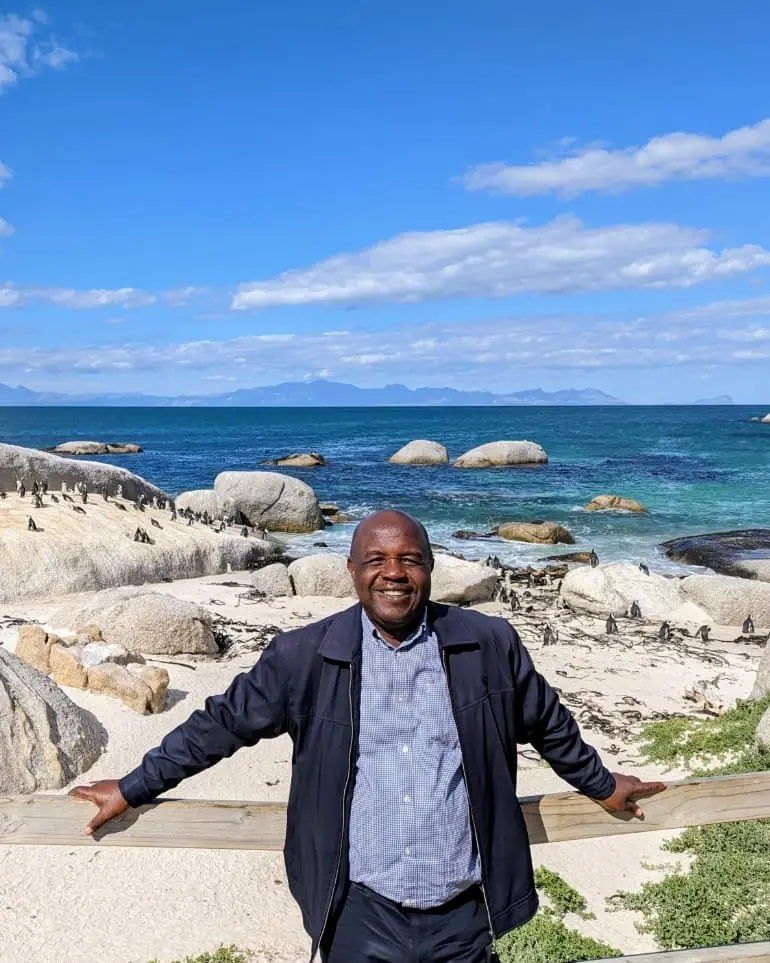 Dr. Peter Yuh Kimeng smiling at a wooden rail on a beach, with penguins on the boulders and the sea behind him
