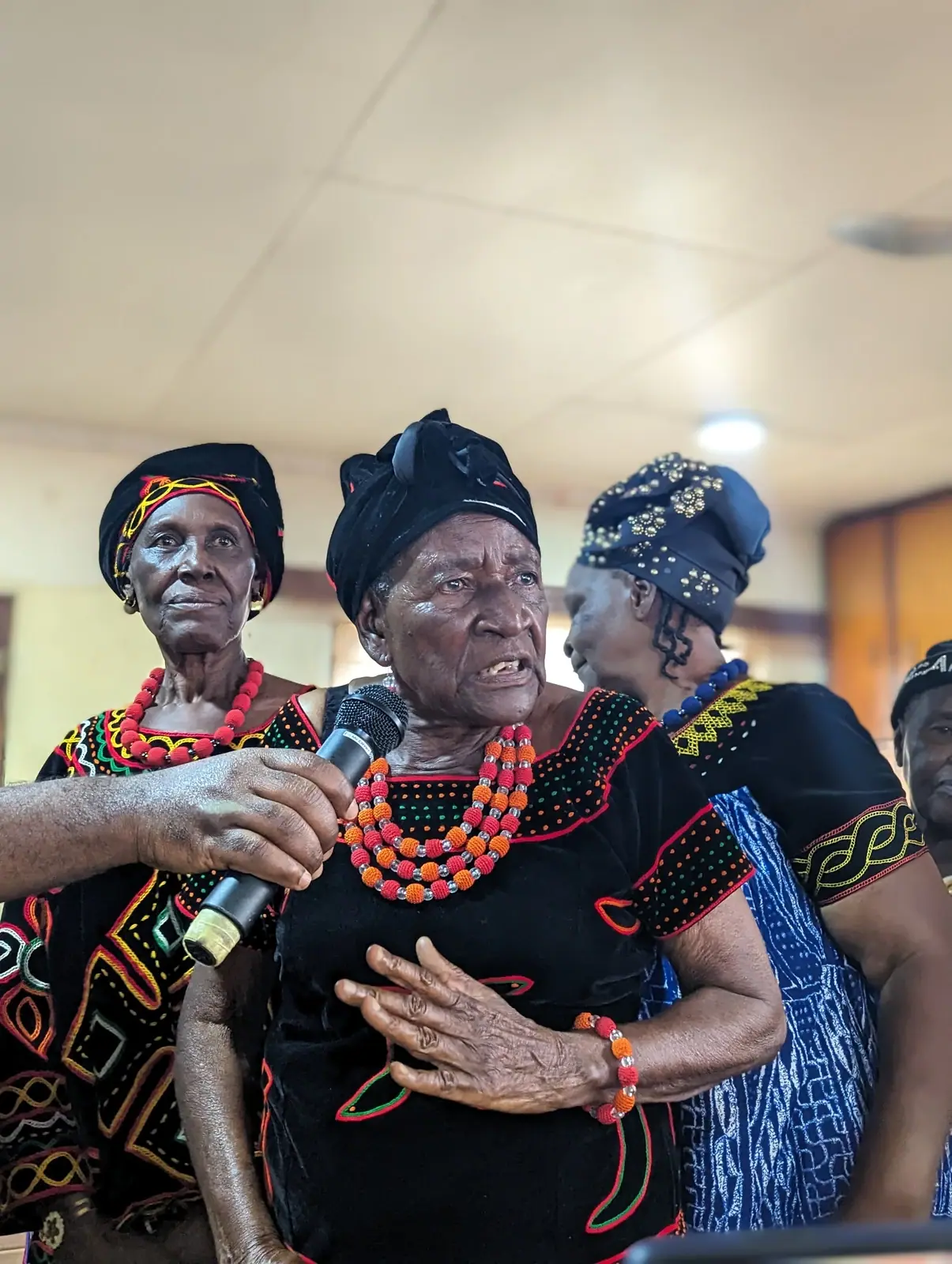 A Kom grandmother in Toghu cloth speaks into a microphone at the dedication of the booklet, two women beside her